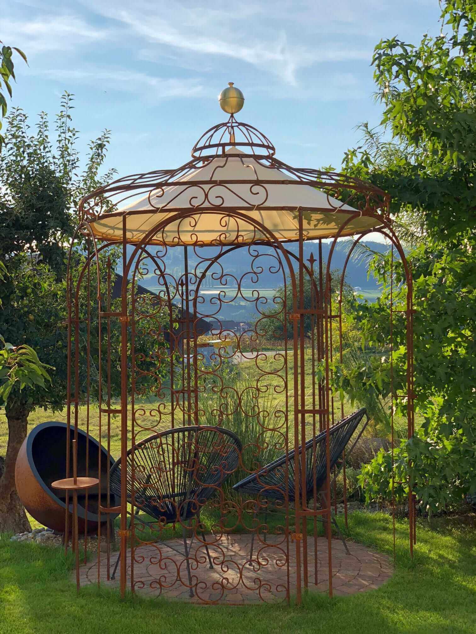 Gartenpavillon Siena mit Sonnensegel und Rankgitter Rosa in einem Garten mit zwei Stuehlen