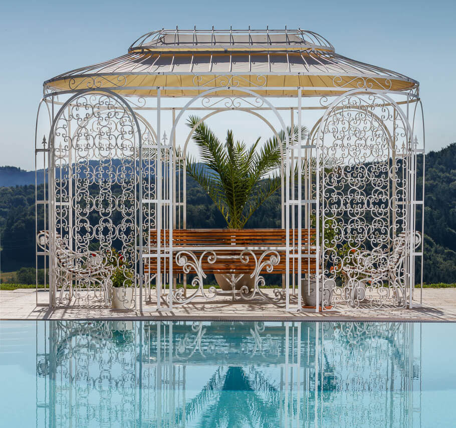 Pavillon Verona in pulverbeschichteter Sonderfarbe weiss mit Sonnensegel und Rankgitter hinter modernen Pool